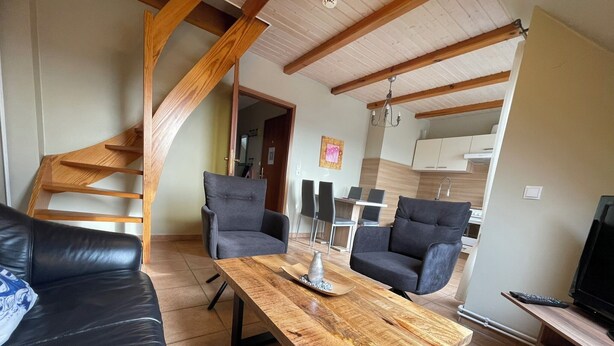 A living room with a wooden coffee table, two grey armchairs, a black leather sofa, a TV, and a staircase leading to an upper floor.