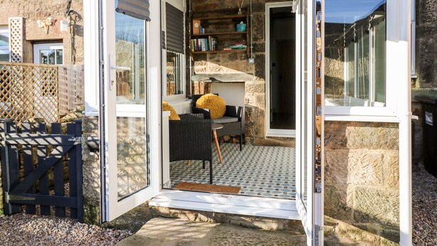 Une porte ouverte donne sur une pièce dont le sol est recouvert de carreaux, où l’on trouve une table avec des chaises et une bibliothèque.