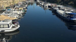 A marina with boats docked along a river, a bridge in the background, and buildings on the opposite bank.