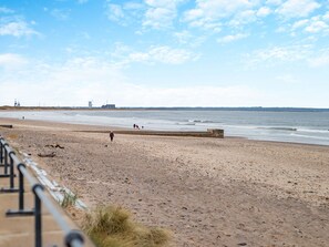 Ein sandiger Strand mit einer Betonbarriere, ein paar Spaziergänger und Industriegebäude in der Ferne.