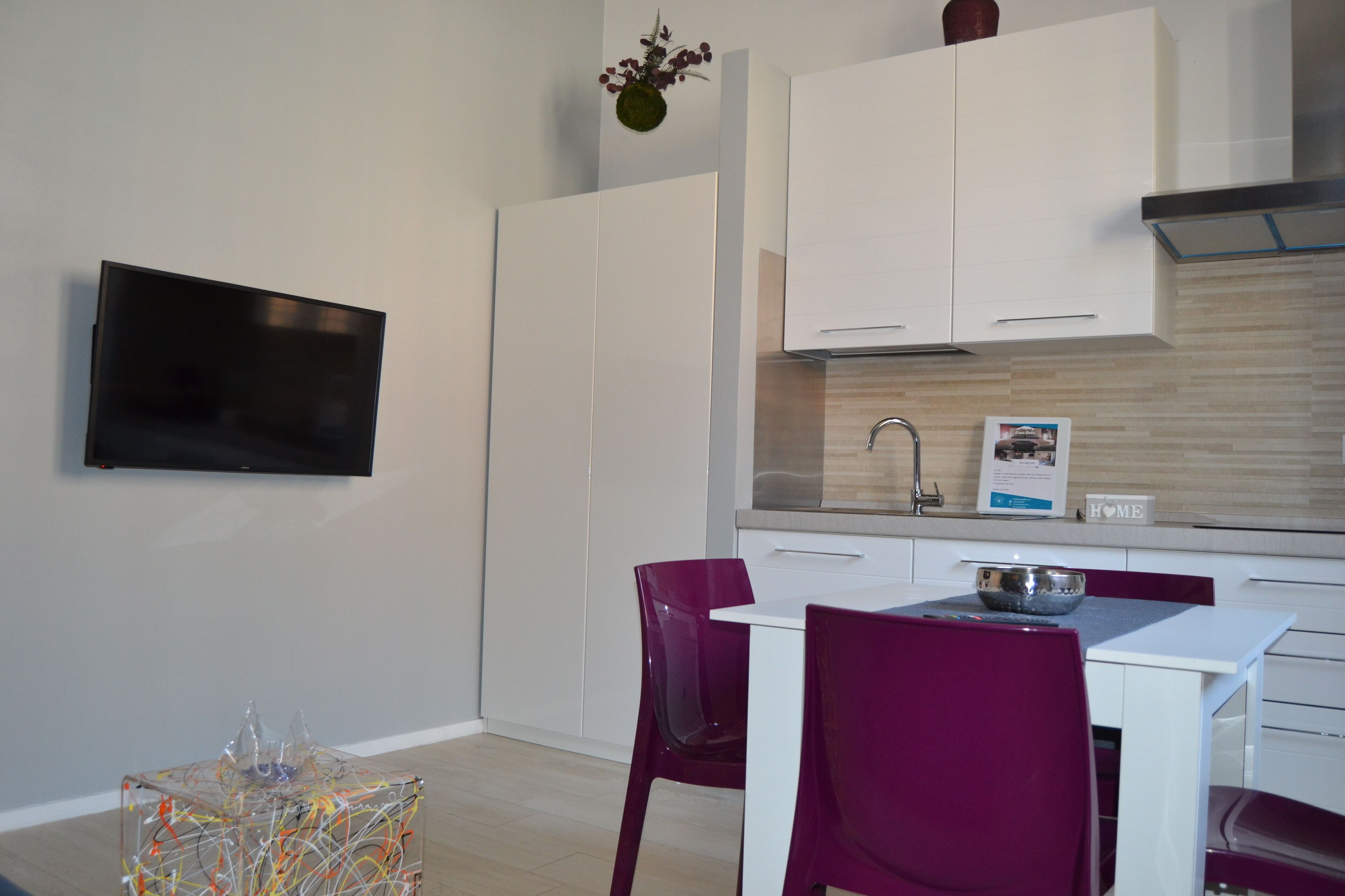 A modern kitchen with white cabinets, a built-in oven, and a stainless steel sink. There is a dining table with purple chairs and a small decorative plant on the wall.