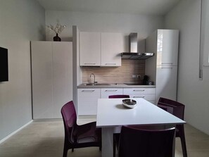 Una cocina moderna con armarios blancos, un lavamanos de acero inoxidable y un salpicadero de madera. Hay una mesa de comedor con dos sillas y un florero sobre un mueble.