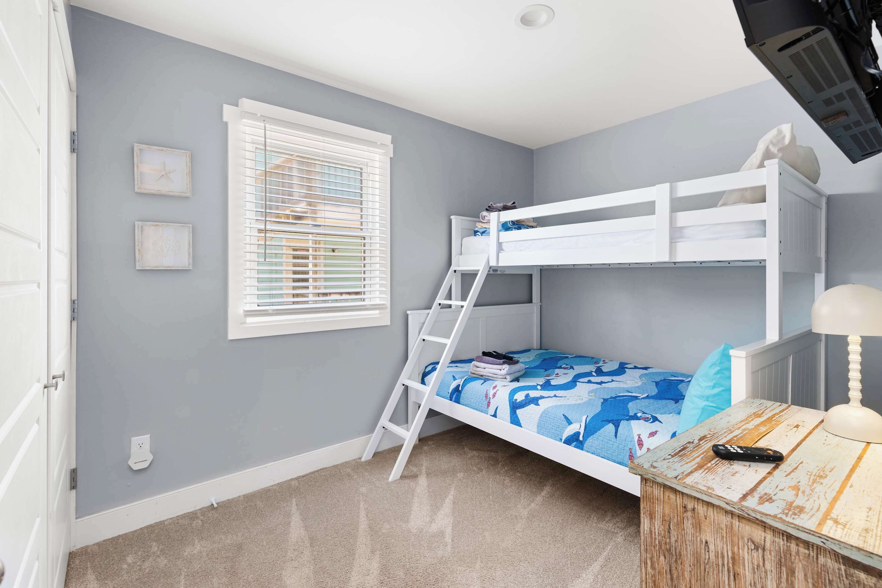 Een stapelbed met een ladder, een raam met zonwering, een nachtkastje met een lamp en een stopcontact op de muur.