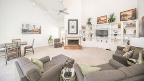 A spacious living room with a fireplace, grey sofas, a dining area with a wooden table and chairs, and a white shelving unit with decorative items and a television.