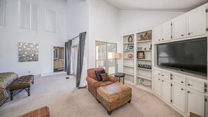 A spacious living room with a large flat-screen TV, a plaid sofa, a matching ottoman, and a bookshelf with decorative items.