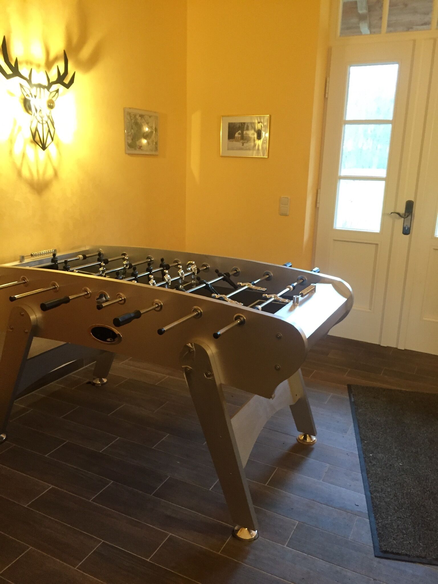 A foosball table in a room with a wooden floor, a door with a window, and two framed pictures on the wall.