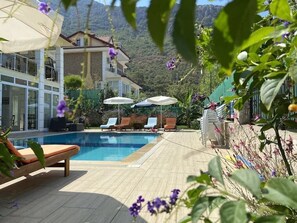 Ein Poolbereich mit Liegestühlen und Sonnenschirmen, umgeben von Grün und mit Blick auf Berge.