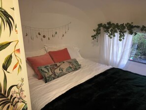 Quarto com cama, almofadas, manta, guirlanda pendurada e janela com planta.