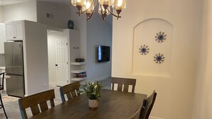 Ein Essbereich mit Holztisch, Stühlen und einem Blumentopf. Es gibt eine Küchenzeile mit Kühlschrank und Fernseher an der Wand.