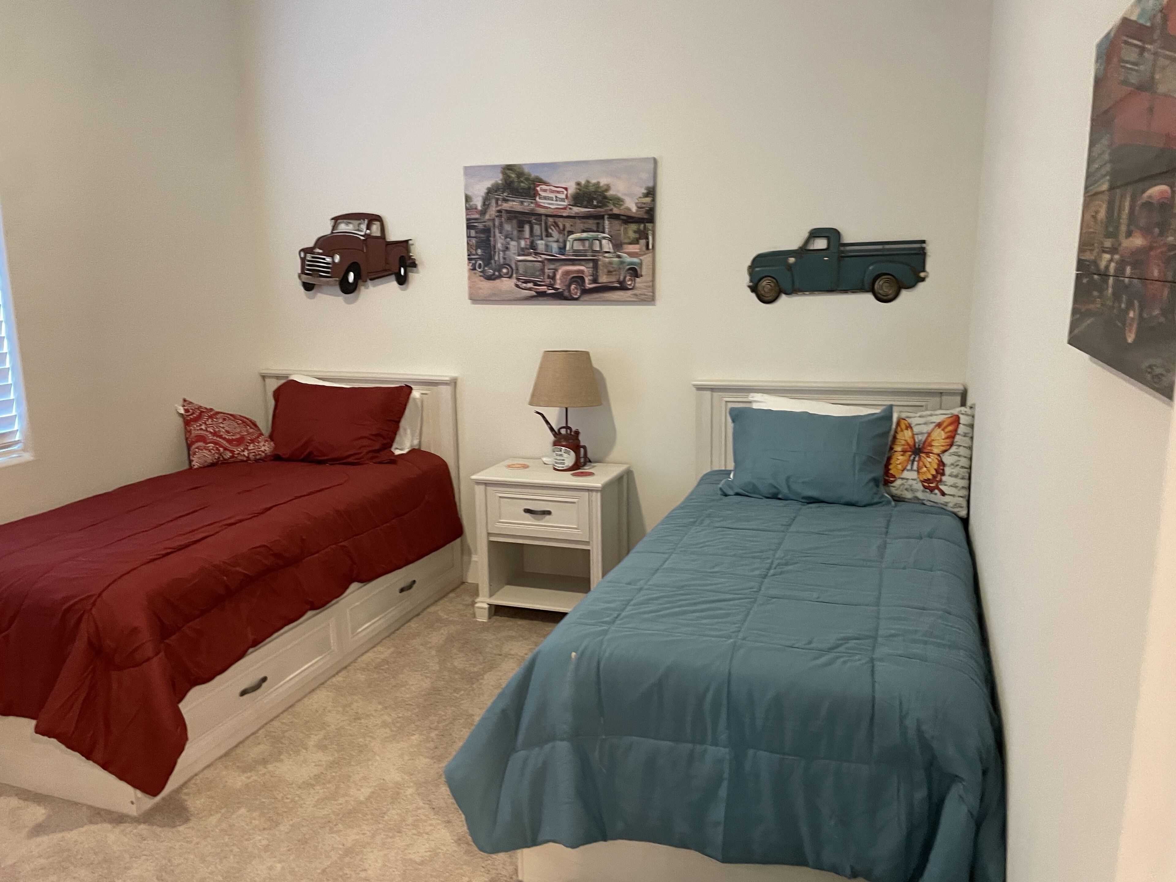 A bedroom with two beds, each with a different colored bedspread, a nightstand with a lamp, and wall decorations including toy trucks and a painting.