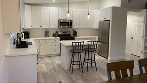 Eine moderne Küche mit weißen Schränken, einem Edelstahl-Kühlschrank, einer Mikrowelle und einem Gasherd. Zwei Barhocker und ein Holztisch sind ebenfalls vorhanden.