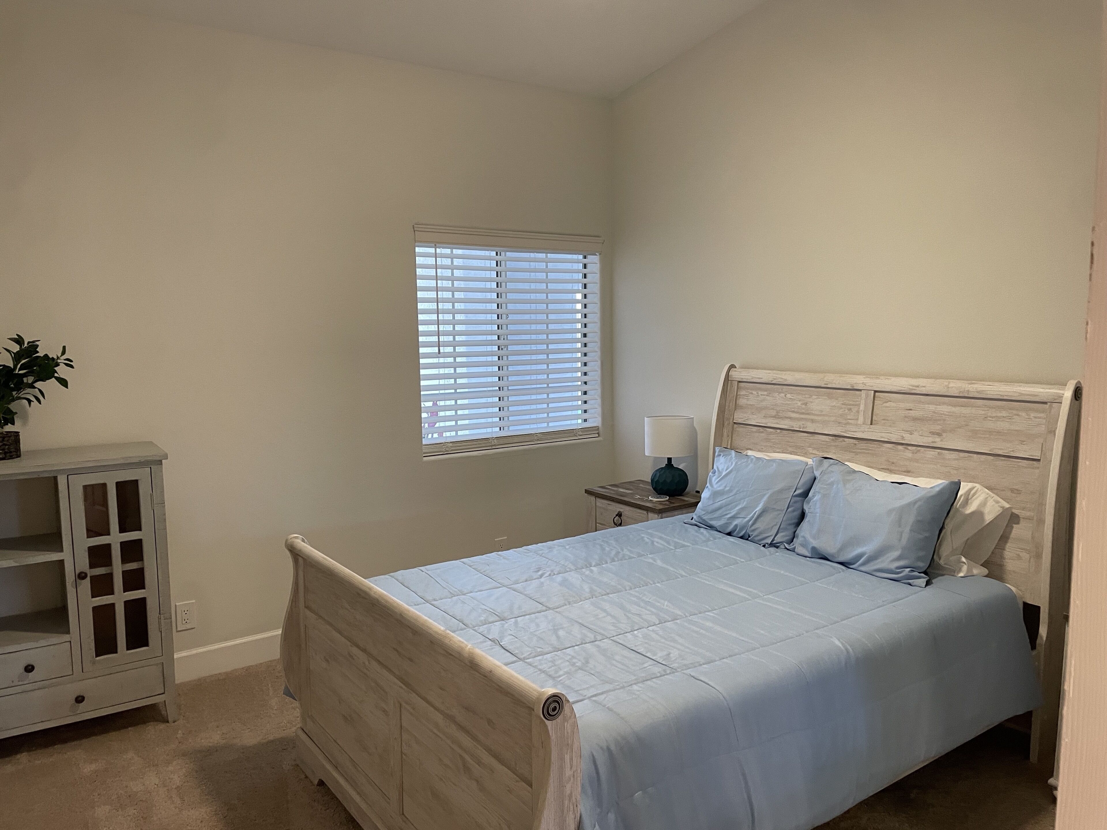 A bedroom with a bed, a nightstand, a window with blinds, and a small cabinet.