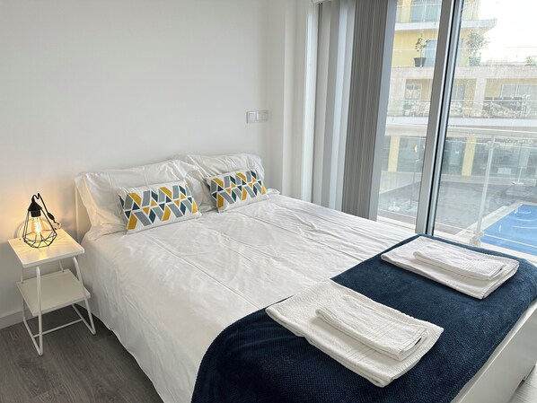 Habitación de hotel con cama, almohadas, mesita de noche, lámpara y vistas al exterior.