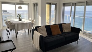 A modern living room with a dark sofa, a round table, and chairs. Large windows offer a view of the ocean. - A-MO Sesimbra by Trip2Portugal (Sesimbra)
