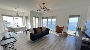 A modern living room with a black sofa, a dining table with chairs, a ceiling light fixture, and large windows offering a view of the ocean. - A-MO Sesimbra by Trip2Portugal (Sesimbra)
