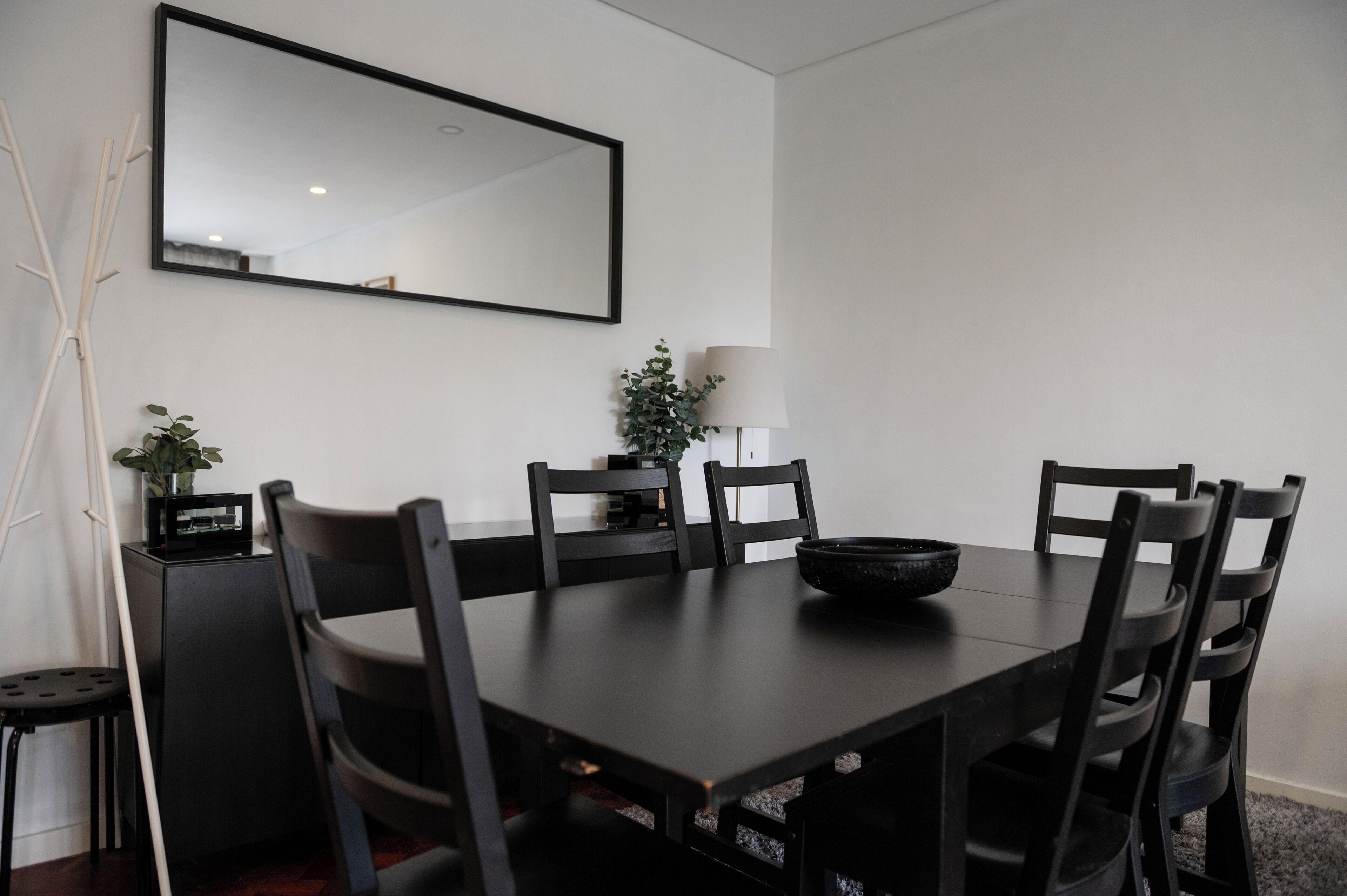 A dining area with a rectangular table, six chairs, a black bowl, a potted plant, a lamp, and a mirror on the wall.