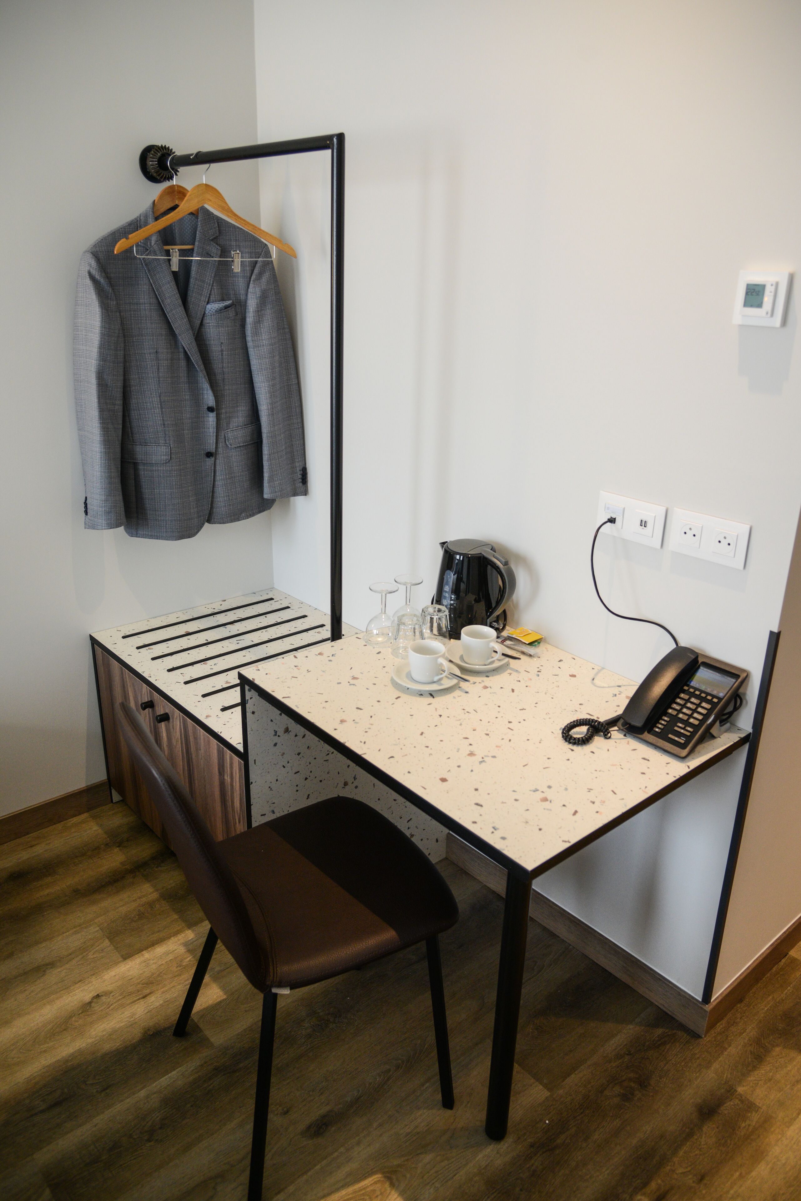 A compact hotel room with a small desk, a chair, a coat rack with a suit, a coffee maker, and a phone.