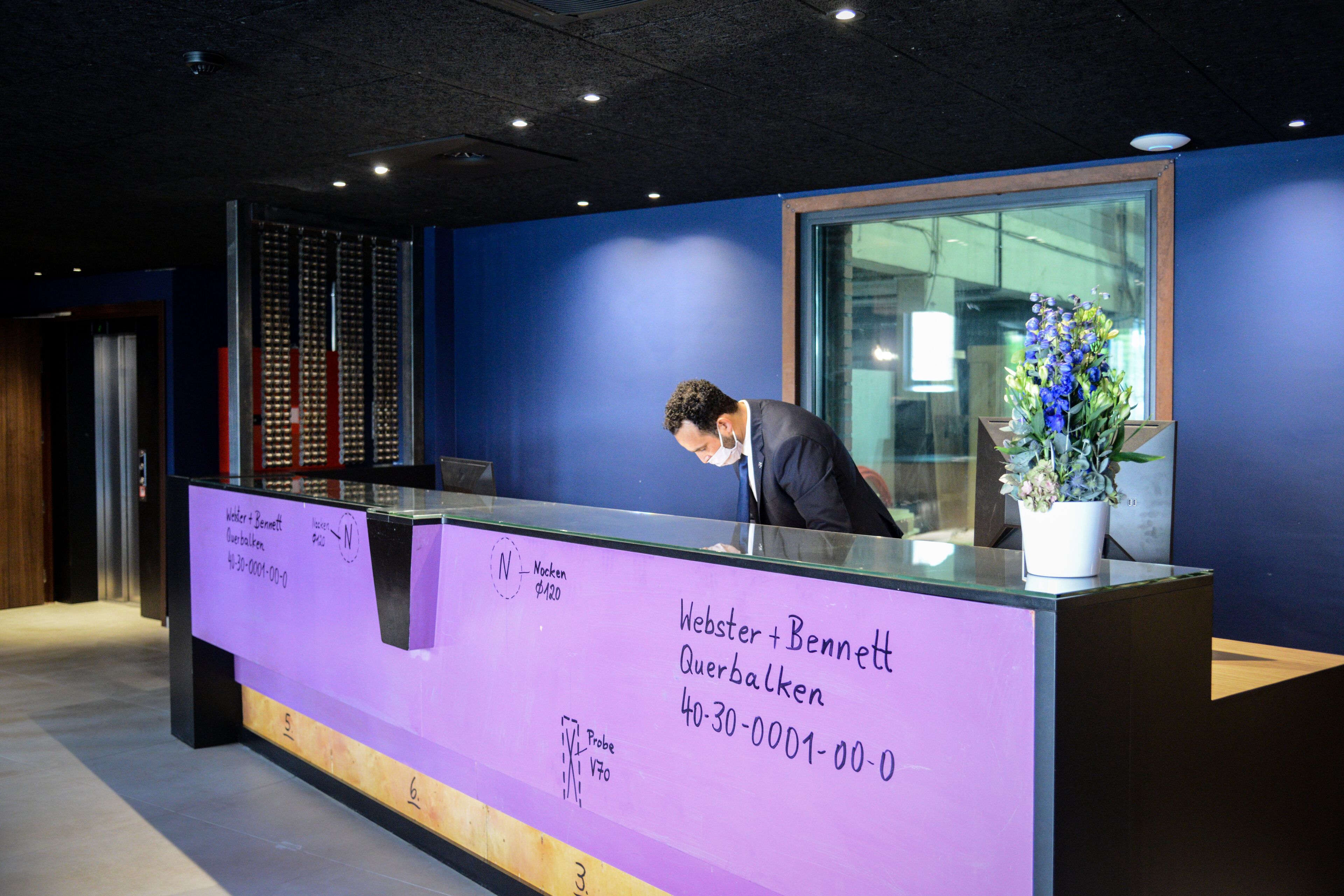 A reception desk with a purple sign, a potted plant, and a glass partition.