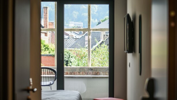 A narrow hallway leading to a bedroom with a large window, a bed, a red ottoman, and a chair. - Pegard Andenne Centre (Andenne)