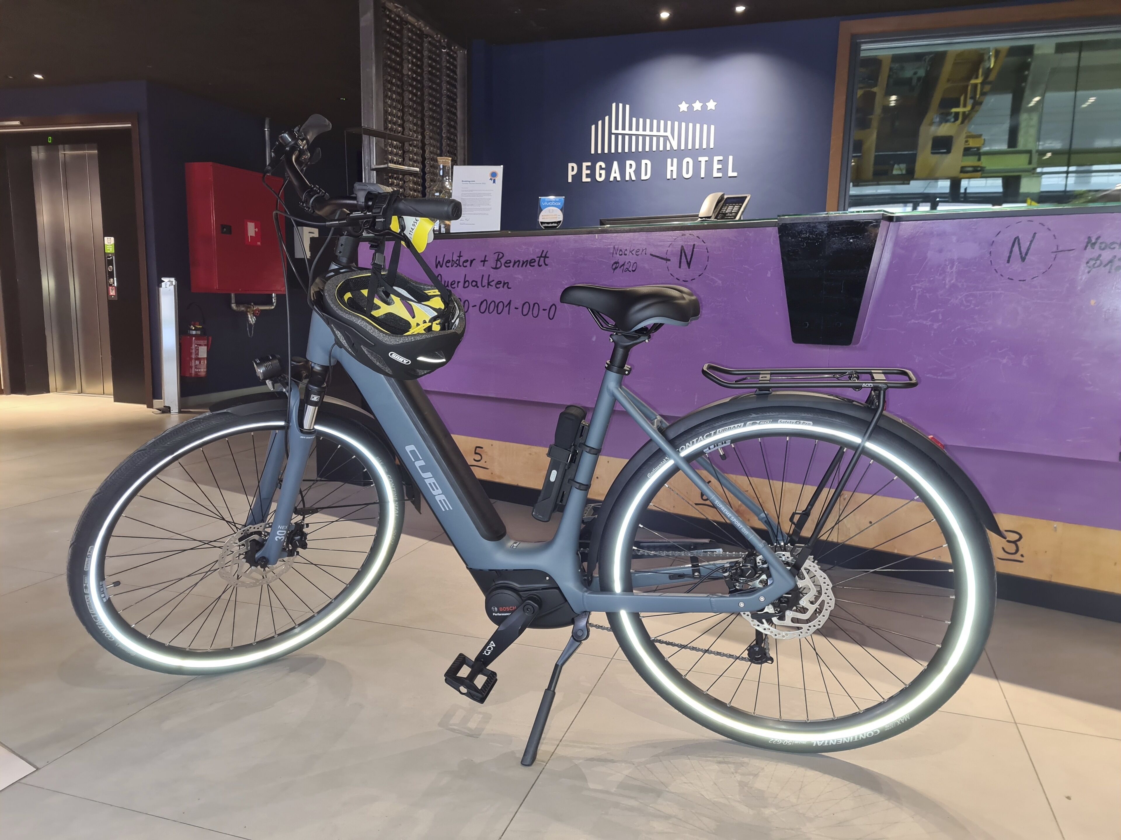 Un vélo avec une selle noire et un panier à l’avant, garé devant le comptoir de la réception d’un hôtel dont le mur est violet et sur lequel on peut lire l’inscription « HÔTEL PEGARD ».