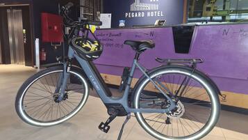 Un vélo avec une selle noire et un panier à l’avant, garé devant le comptoir de la réception d’un hôtel dont le mur est violet et sur lequel on peut lire l’inscription « HÔTEL PEGARD ».