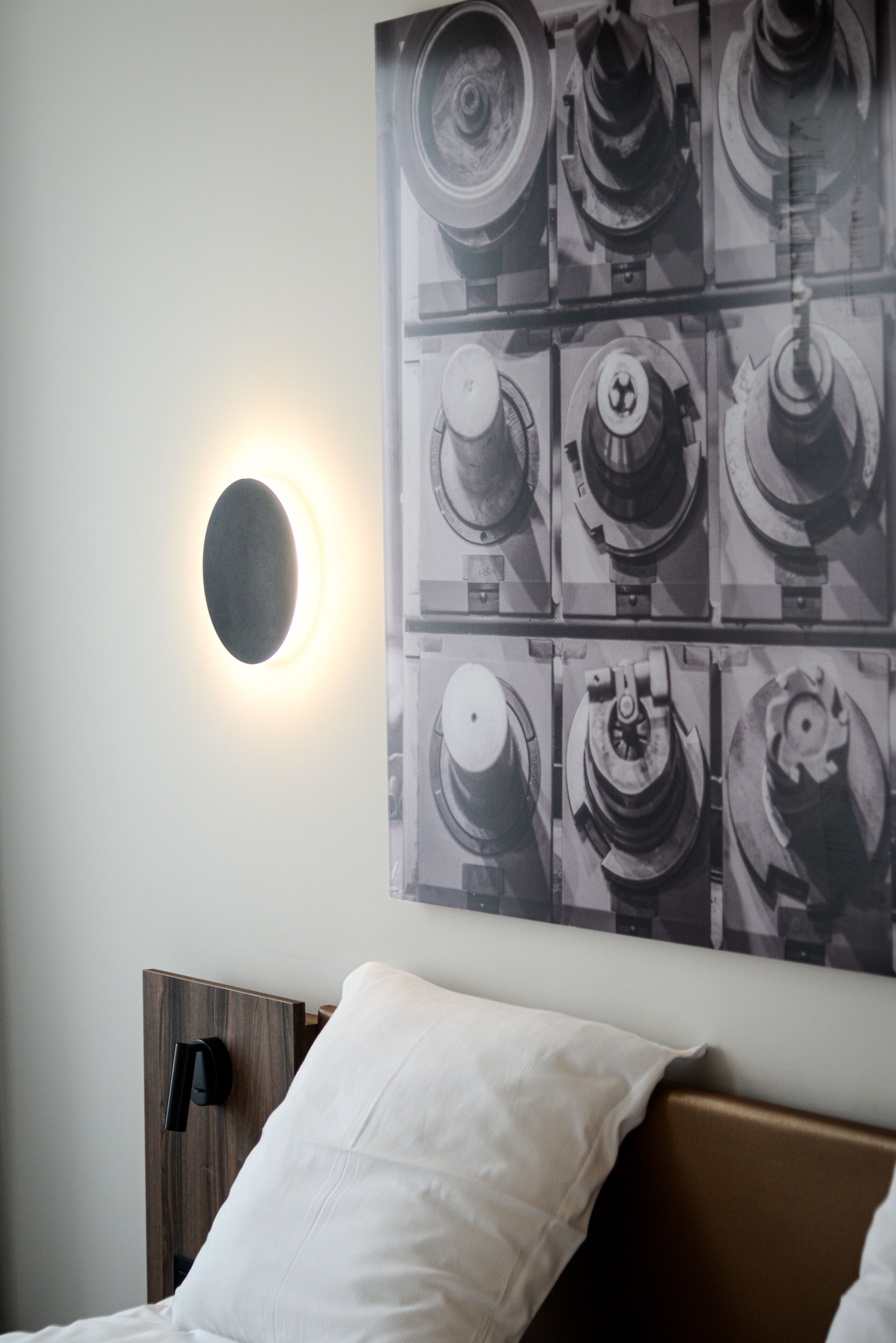 A bedroom with a bed, a headboard, a wall-mounted light, and a poster of mechanical parts.