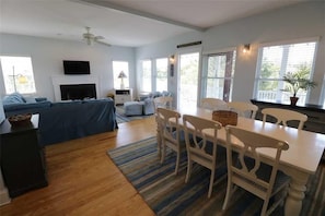 A spacious living room with a sofa, dining table, and chairs. There is a fireplace, a television, and a ceiling fan.