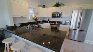 A modern kitchen with white cabinets, a stainless steel refrigerator, a microwave, and a stove. There are three white stools at the island. - Oceanview cottage steps to beach access and close to Marina, BHI (Bald Head Island)