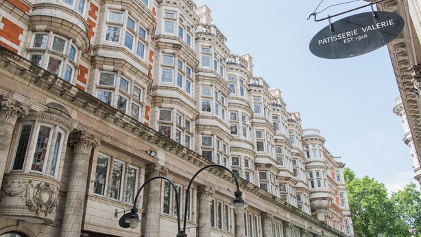 Um edifício histórico com arquitetura elaborada, uma placa com o nome 'Patisserie Valerie' pendurada no canto e um poste de iluminação na frente.
