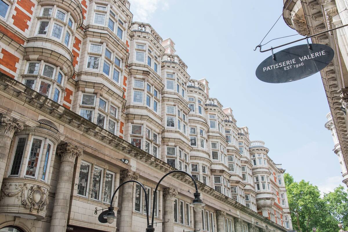 Ein historisches Gebäude mit aufwendiger Architektur, eine Schilder mit dem Schriftzug 'Patisserie Valerie' hängt in der Ecke, und eine Straßenlaterne davor.