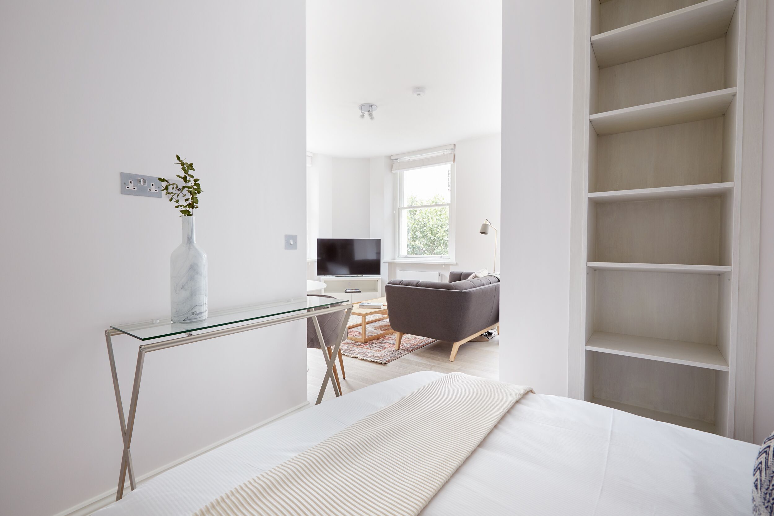 A modern bedroom with a glass desk, a bed with a white comforter, a grey sofa, and a large white shelving unit.