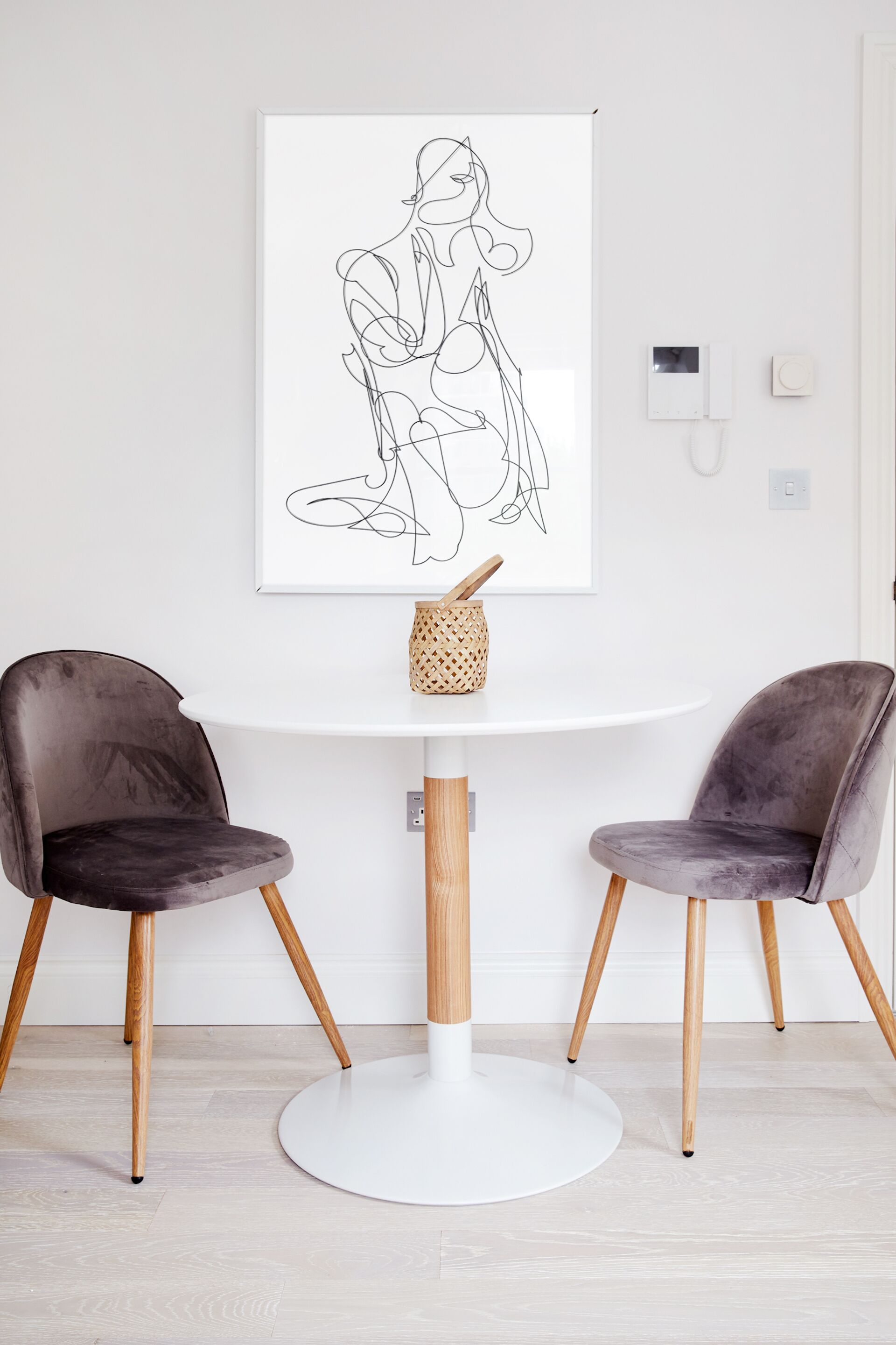 Una habitación moderna con una mesa redonda blanca, dos sillas oscuras y una obra de arte abstracta enmarcada en la pared.