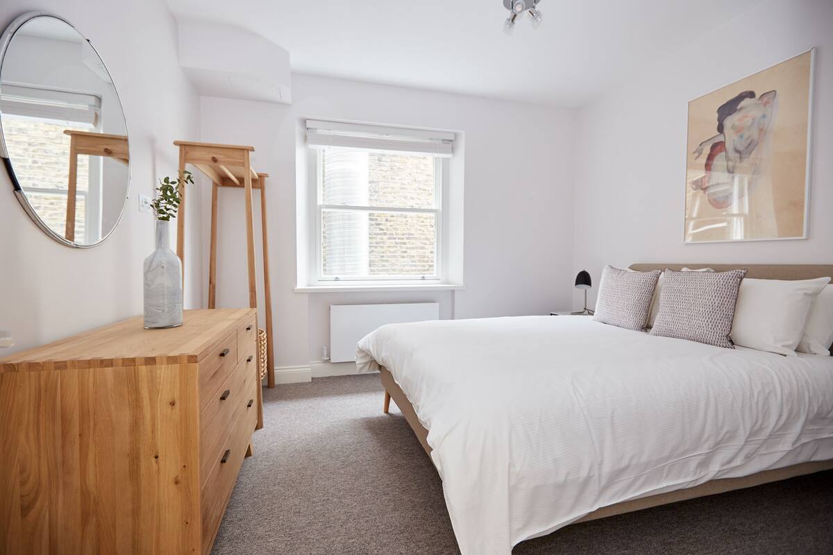 Ein Schlafzimmer mit einem hölzernen Schminktisch, einem Bett mit weißer Bettwäsche, einem großen Spiegel und einem Fenster mit Jalousien.