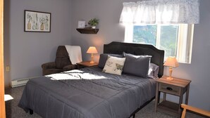 Une chambre à coucher comprenant un lit, deux tables de chevet, une chaise, une fenêtre avec un rideau en dentelle et une étagère murale ornée d’une plante et d’un panneau.