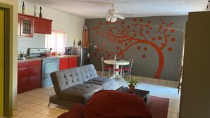 Un espace de vie compact comprenant un coin cuisine, un canapé gris, une table à manger avec des chaises et une fresque murale représentant un arbre décoratif.