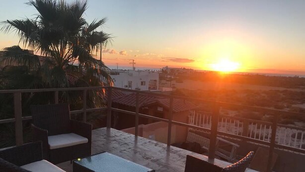 Un balcon doté de meubles en osier et offrant une vue sur le coucher de soleil au-dessus d’un paysage urbain.