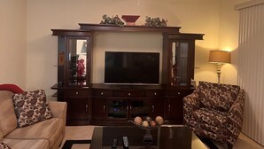 Una sala de estar con televisor de pantalla plana, mueble de madera oscura para el equipo de entretenimiento, dos sillones estampados, una mesa de centro con adornos y un ventilador de techo.