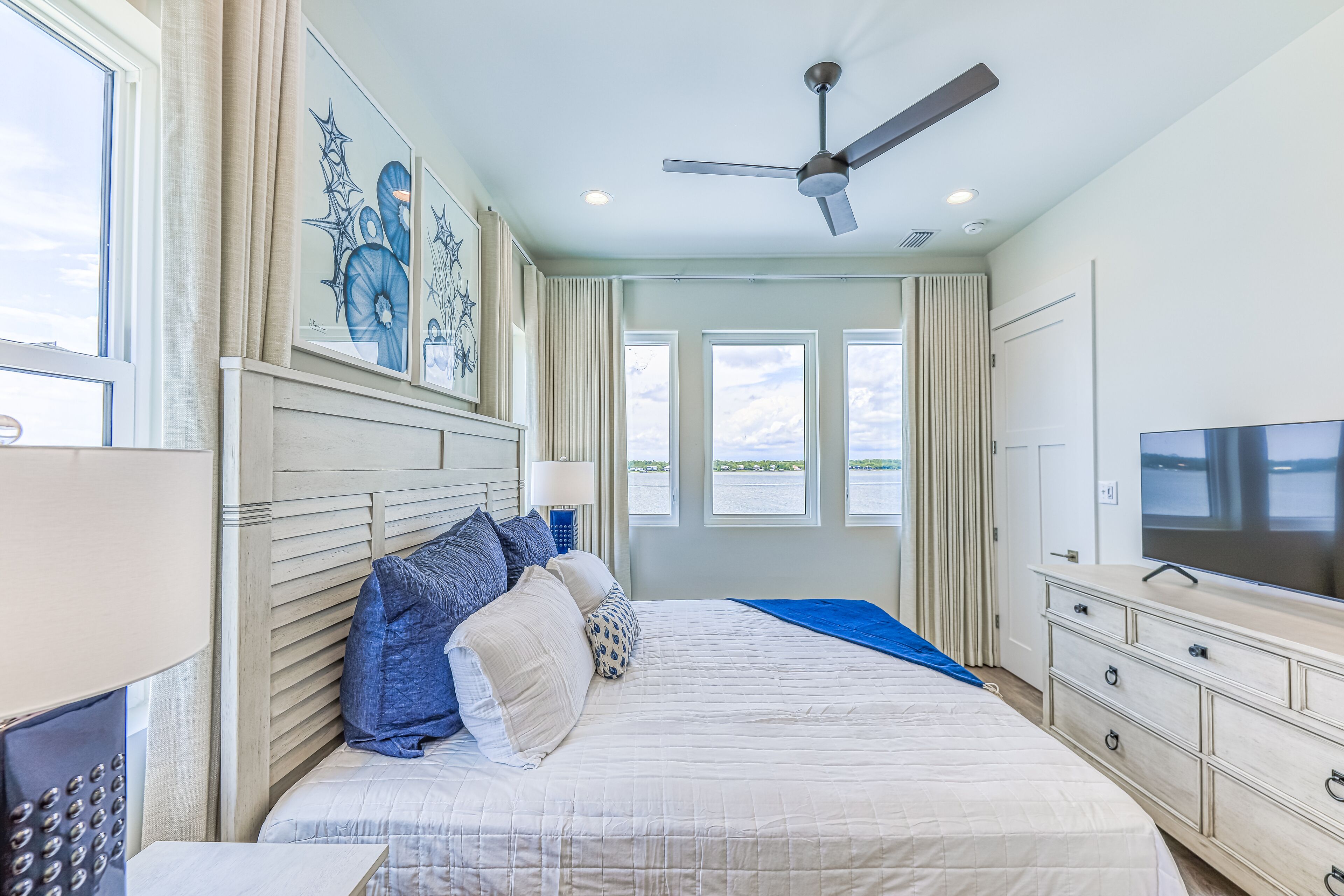 A bedroom with a large bed, a ceiling fan, a TV, and a view of the water outside.