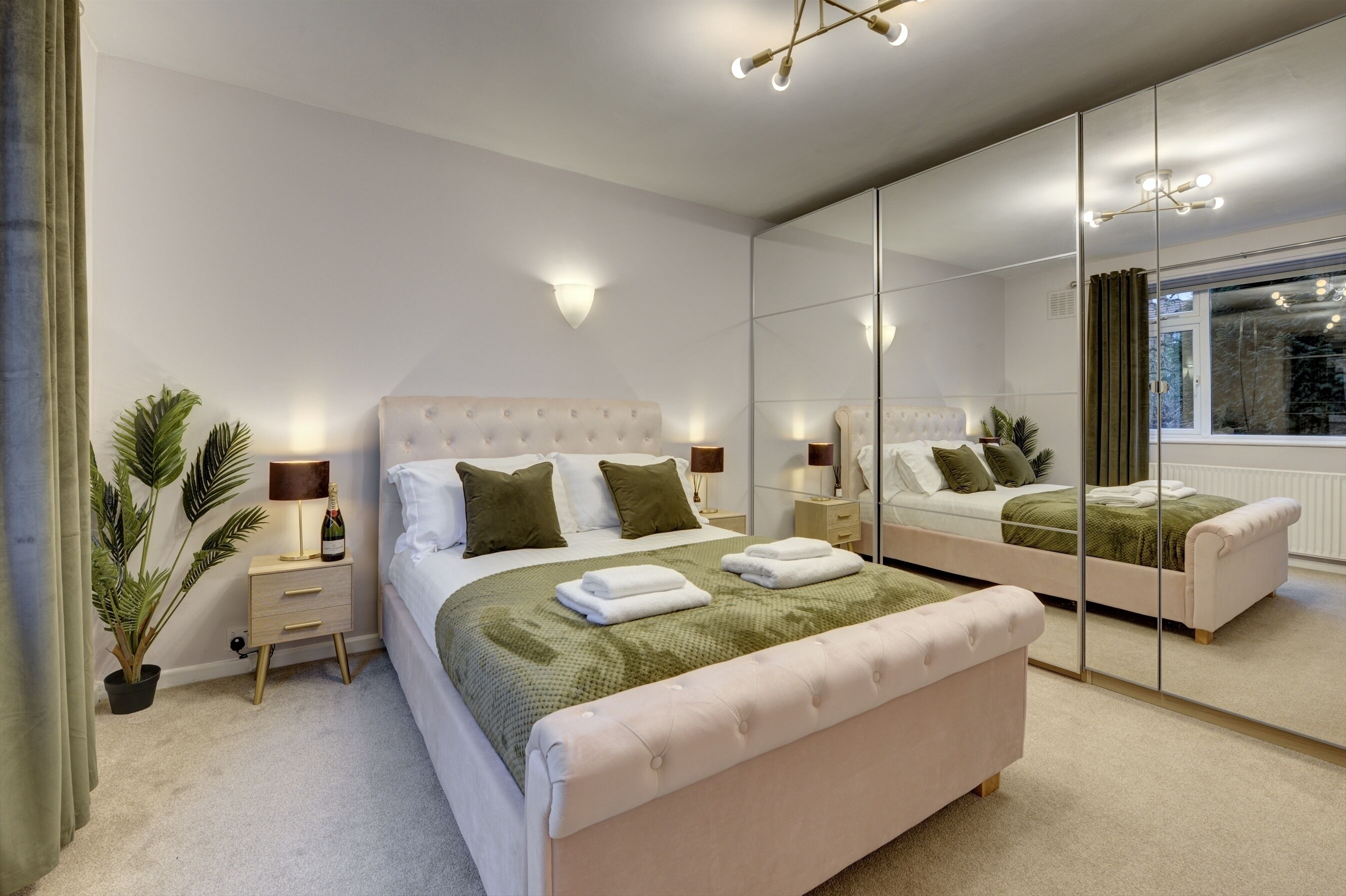 A modern bedroom with a large mirror, a bed with green and white bedding, a nightstand with a lamp, and a potted plant.