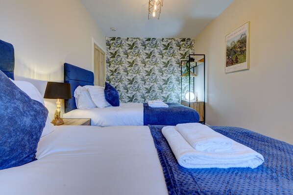 A hotel room with two beds, a large window, a wall with a leafy pattern, and a framed picture on the wall.