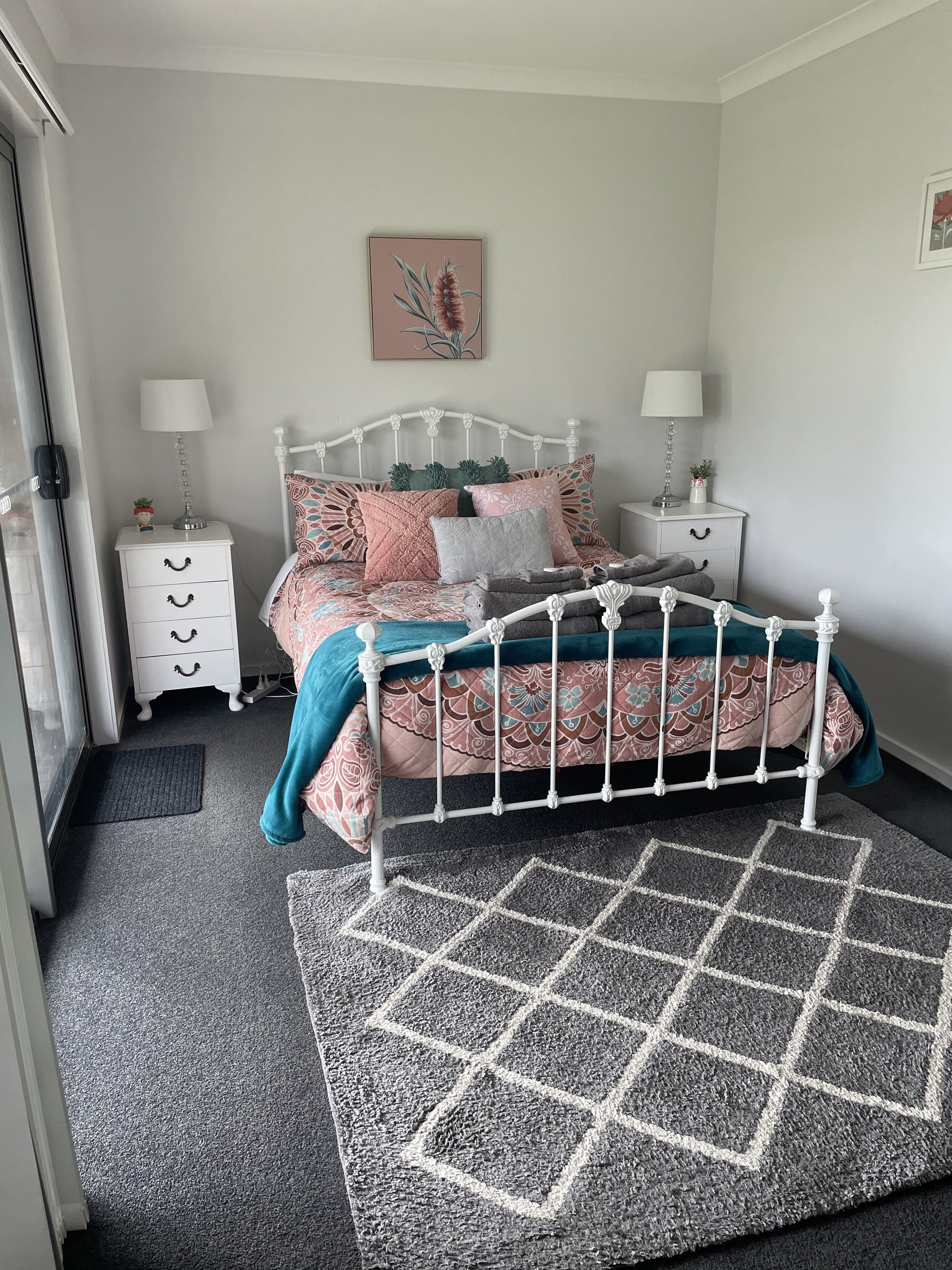 Un dormitorio con estructura de cama metálica blanca, ropa de cama estampada, dos mesitas de noche con lámparas, una planta pequeña en maceta y un adorno en la pared.