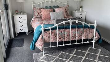 Un dormitorio con estructura de cama metálica blanca, ropa de cama estampada, dos mesitas de noche con lámparas, una planta pequeña en maceta y un adorno en la pared.