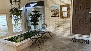 Un área de recepción con suelo de piedra, un mostrador de recepción, dos sillas, una planta colgante con hiedra, una cámara de seguridad montada en la pared, una puerta con un diseño geométrico y un felpudo.