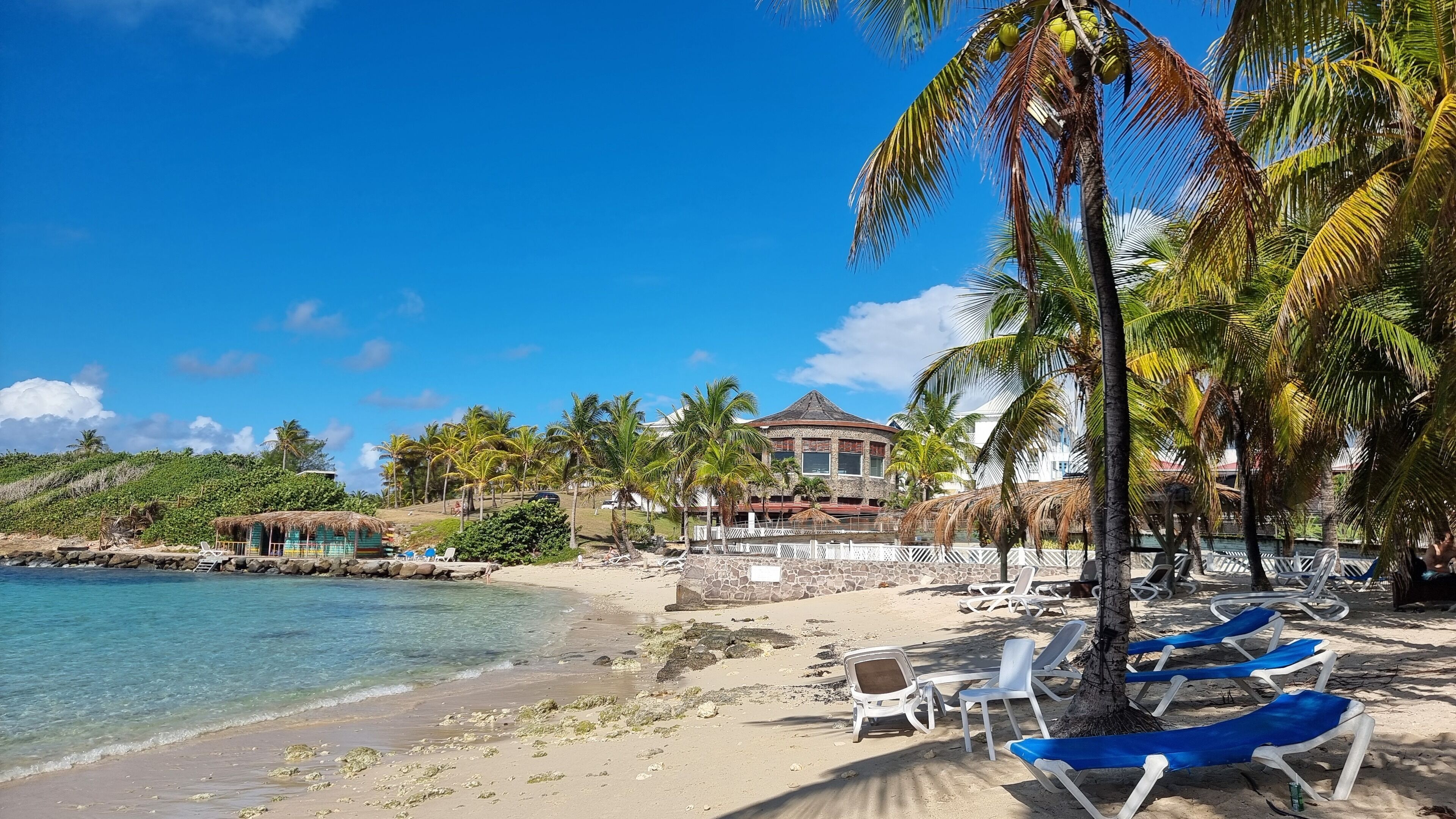 A beach with clear blue water, a thatched-roof hut, and a large house with a stone facade.