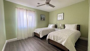 A bedroom with two single beds, a ceiling fan, a nightstand with a lamp, and a window with sheer curtains.