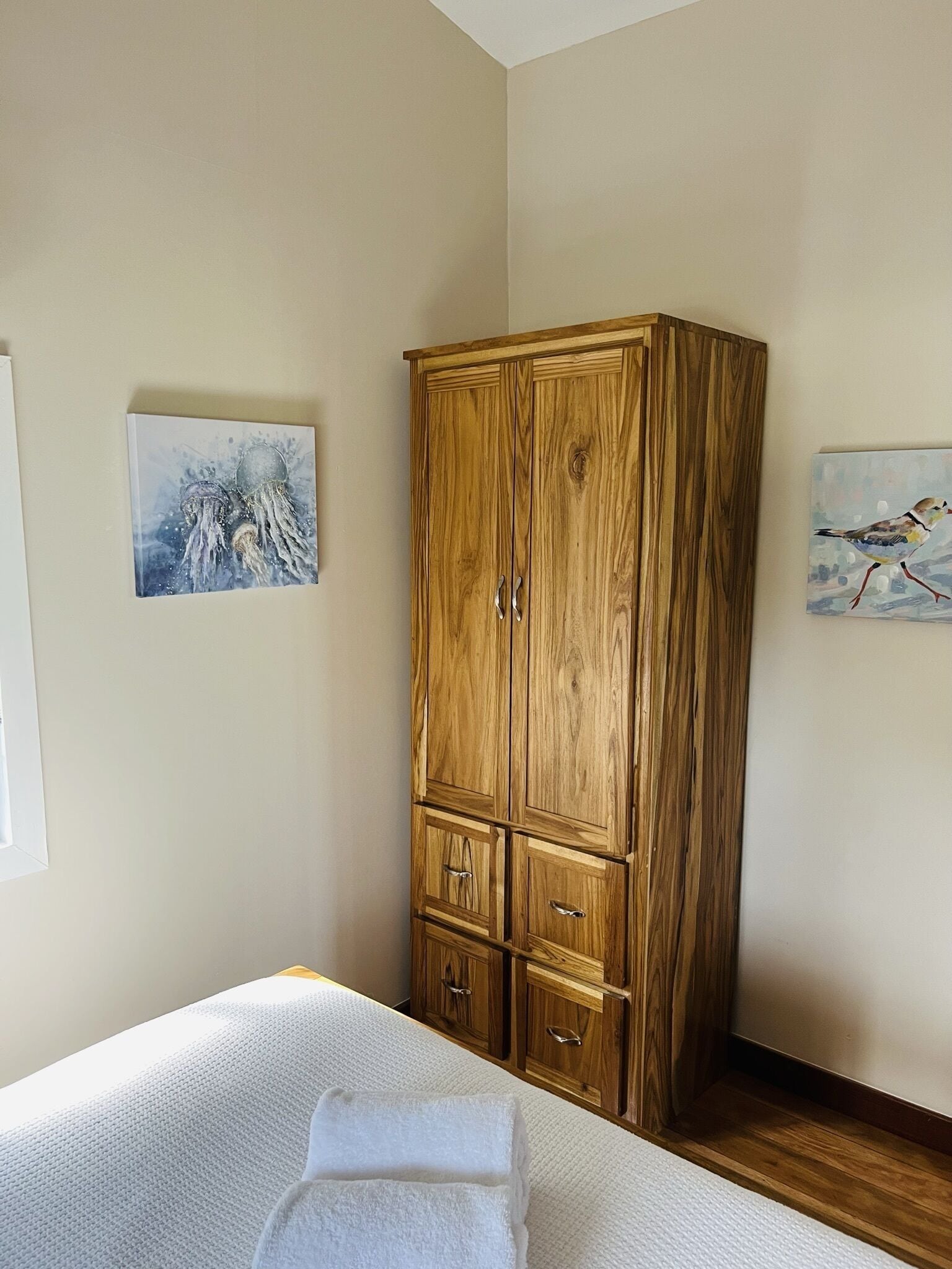 A bedroom with a wooden wardrobe, a bed with a white duvet, and two paintings on the walls.