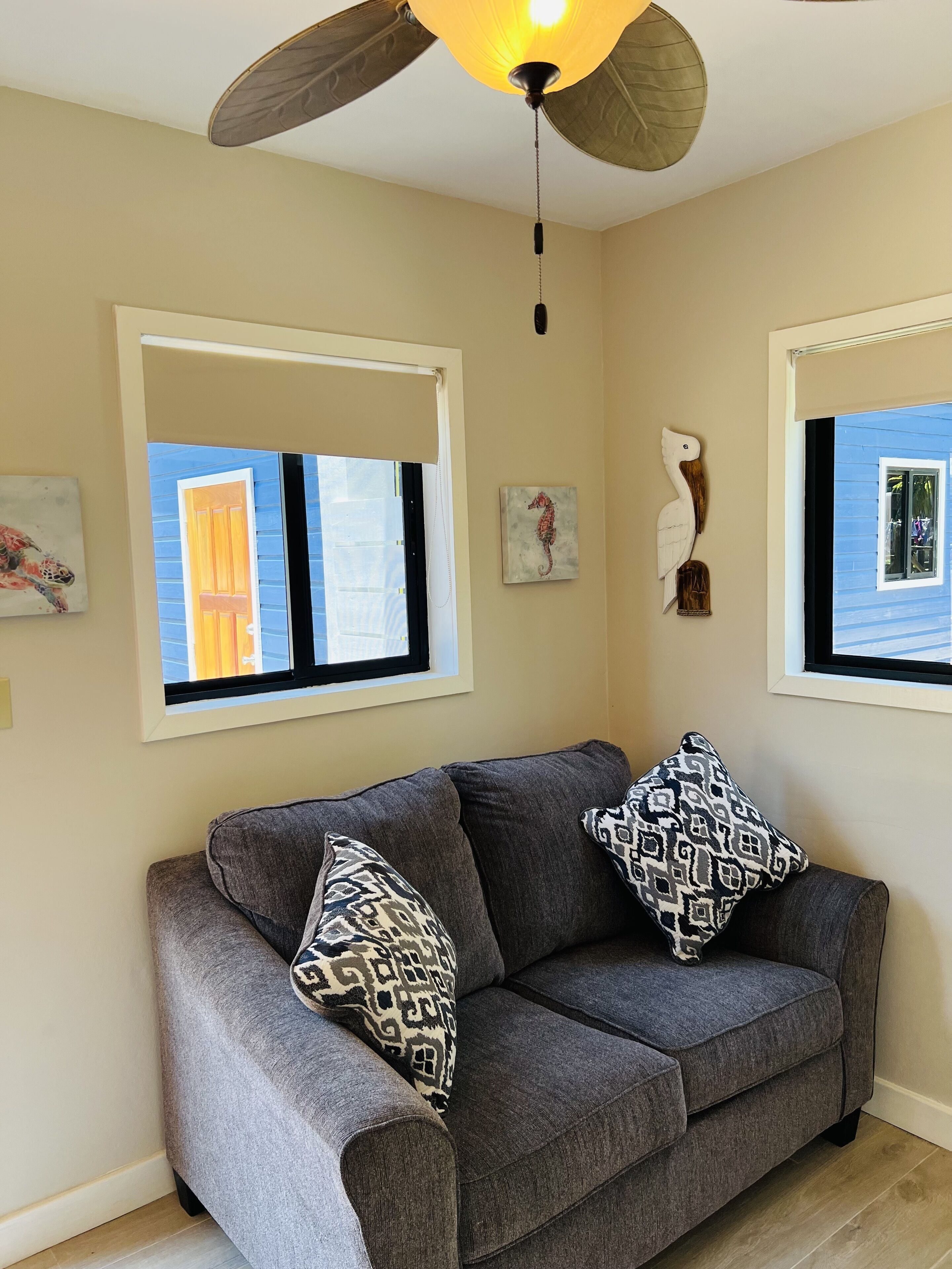A living room with a grey sofa, patterned cushions, a ceiling fan, and windows with blinds.