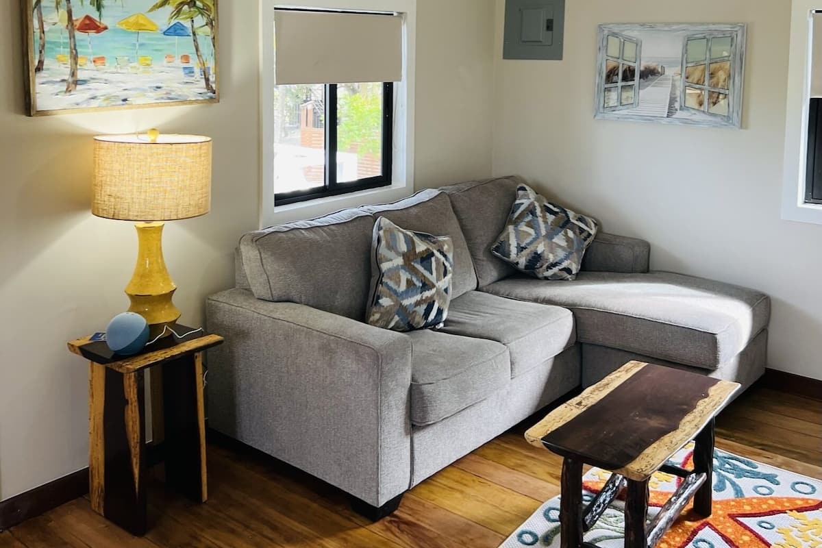 A cozy living room with a grey sofa, a small wooden side table with a lamp, and a colorful rug.