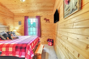 A wooden cabin room with a bed, a bench, a window with curtains, a mounted television, and a wall-mounted sign.