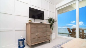 Ein modernes Wohnzimmer mit einem Holzschrank, einem Flachbildfernseher, einem Topf mit Pflanze und Blick auf den Ozean durch große Glasfenster.